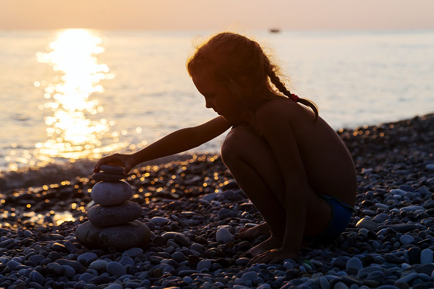 Silhouette of little young girl on sea, puts a pyramid from stones  at the sunset